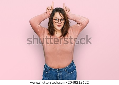 Young plus size woman wearing casual clothes and glasses doing funny gesture with finger over head as bull horns 