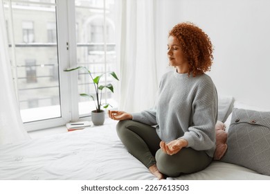 Young plus-size redhead woman with fluffy curly hair sitting in lotus posture on bed near big window keeping hands on knees, meditating, trying to relax her mind and relieve stress - Powered by Shutterstock