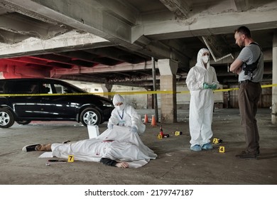 Young Policeman Communicating With Forensic Expert At Crime Scene With Dead Body Covered With White Sheet In Parking Area