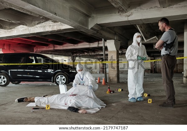 Young\
policeman communicating with forensic expert at crime scene with\
dead body covered with white sheet in parking\
area