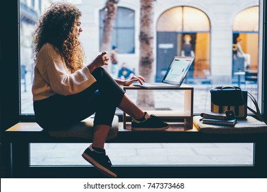 Young pondering female person with beautiful curly hair checking account balance on webpage.Talented student preparing for upcoming examinations during sitting next to copy space for your advertising