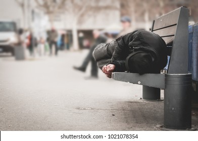 Young poor dirty homeless man or refugee sleeping on the wooden bench on the urban street in the city, social documentary concept