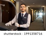 Young porter in uniform standing by cart in long corridor of modern hotel