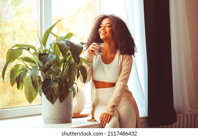 Young positive african american woman sitting on windowsill with glass of pure mineral water and looking through window with smile, drinking aqua in morning, starting new day with healthy habits - Shutterstock ID 2117486432