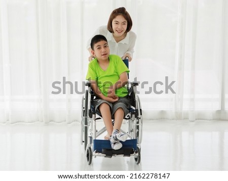 Young positive Asian female caregiver rolling wheelchair with adorable smiling ethnic boy living with cerebral palsy disability in modern clinic