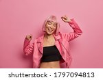 Young positive Asian woman with bob hairstyle, dances and has fun, keeps arms raised, wears black tank top and rosy jacket, moves to music, has happy look, closes eyes in joy, pink background