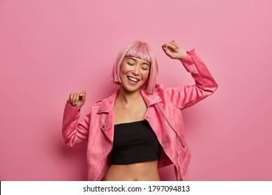 Young positive Asian woman with bob hairstyle, dances and has fun, keeps arms raised, wears black tank top and rosy jacket, moves to music, has happy look, closes eyes in joy, pink background - Powered by Shutterstock