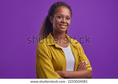 Young positive beautiful African American woman arms crossed looking at camera posing in confident and self-sufficient pose human dressed in casual oversize clothes stands in purple studio