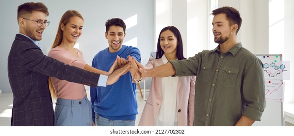 Young Positive Business Team Fold Hands Together As Symbol Of Cooperation, Unity And Success. Joyful Smart Men And Women Are Set For Successful Outcome. Human Support Concept. Banner.