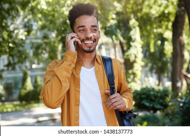 Young positive dark skinned man in yellow shirt, walking at the park, speaking on smartphone, waitng his friend, looking away and broadly smiling.