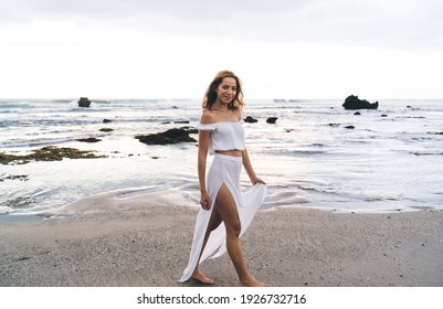 Young positive female with brown hair in white top and slit skirt walking on sandy beach with rocks in water behind