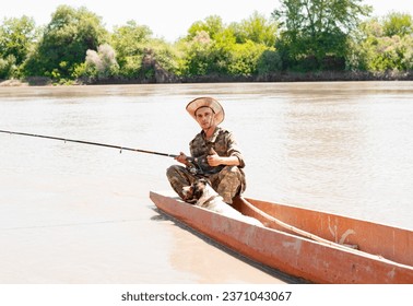 Young, positive fisherman showing thumb up, while sitting on stern boats and fishing. Front view of man with dog gesturing like sign, while holding fishing rod on lake. Concept of positive emotions. - Powered by Shutterstock