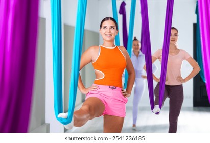 Young positive girl practicing aerial yoga with group of women in fitness center, doing exercises with hanging hammock - Powered by Shutterstock