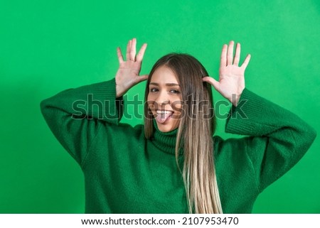 Young positive happy smiling woman making a face with hands and tongue out against green not isolated background. Cheerful female goofing, youth culture concept