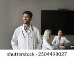 Young positive Indian man doctor wearing white coat, standing in clinic conference room feels happy, looking into distance, thinking, dreaming. Medical worker portrait, mission, occupation, healthcare