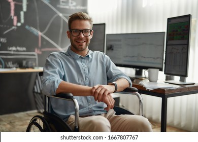 Young And Positive Male Trader In A Wheelchair Smiling At Camera While Sitting At His Workplace In The Modern Office. Disability And Handicap Concept. Handicapped Worker. Trading Online. Stock Market