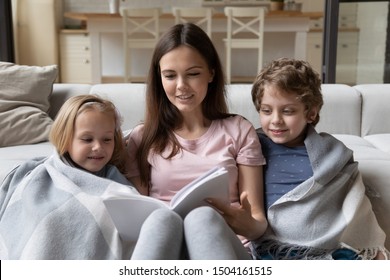 Young Positive Mommy Sitting On Warm Heated Floor With Covered In Plaid Little Adorable Smiling Kids Siblings, Reading Fairy Tale Together. Happy Mother Holding Book, Telling Story To Cute Children.