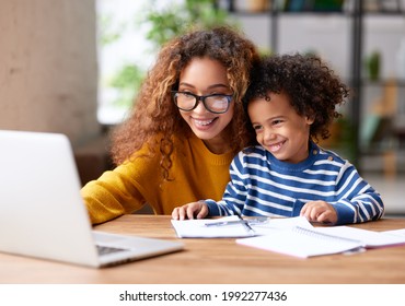 Young positive mother with happy son sitting at desk and usinng laptop, looking at screen and smiling while watching funny videos together. Happy afro american family spending leisure time at home - Powered by Shutterstock