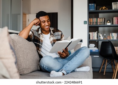Young positive smiling african man wearing plaid shirt reading book while sitting on couch at home