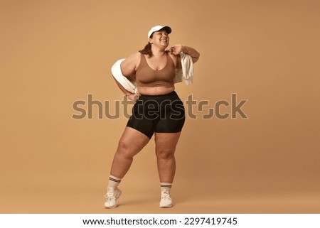 Young, positive, smiling, overweight woman posing in stylish, trendy sportswear with cap against brown studio background. Concept of sport, body-positivity, weight loss, body and health care
