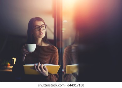 Young positive talented female author reading bestseller and dreaming about personal popular book publication,teen smiling hipster girl having relaxation time standing in library during school break