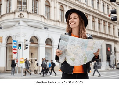 Young positive tourist holds travel paper map, reads route of trip, goes sightseeing, searches destination, gets to right place wears black headgear and leather jacket focused away with pleasant smile