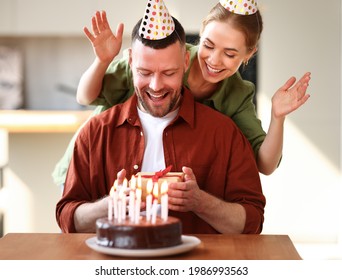 Young Positive Wife Making Surprise For Husband, Excited Man Holding Gift Box And Being So Happy While Sitting In Kitchen At Table With Cake. Family Couple In Party Hats Celebrating Birthday At Home