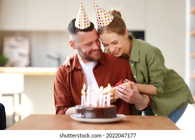 Young Positive Wife Making Surprise For Husband, Excited Man Holding Gift Box And Being So Happy While Sitting In Kitchen At Table With Cake. Family Couple In Party Hats Celebrating Birthday At Home