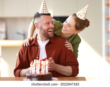 Young Positive Wife Making Surprise For Husband, Excited Man Holding Gift Box And Being So Happy While Sitting In Kitchen At Table With Cake. Family Couple In Party Hats Celebrating Birthday At Home