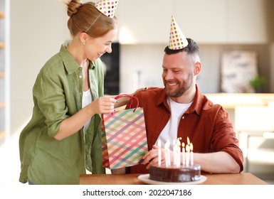 Young Positive Wife Making Surprise For Husband, Excited Man Holding Gift Package And Being So Happy While Sitting In Kitchen At Table With Cake. Family Couple In Party Hats Celebrating Birthday  