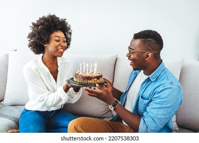 Young Positive Wife Making Surprise For Husband, Excited Man Holding Gift Box And Being So Happy While Sitting In Living Room At Table With Cake. Family Couple In Party Hats Celebrating Birthday