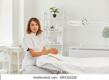 Young positive woman cosmetologist or dermatologist sitting near empty beauticians coach awaiting for client visit and looking at camera in beauty spa salon. Cosmetology and skincare concept