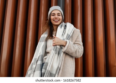 Young positive woman in a fashionable knitted hat in a stylish coat of milk color from eco-fur with a woolen warm scarf poses and cute smiling near a vintage metal wall. Pretty funny girl model. - Powered by Shutterstock