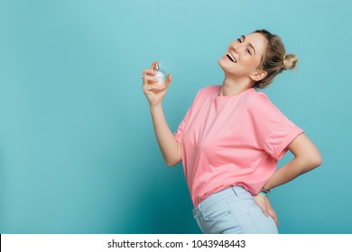 young positive woman holding a perfume bottle and applying it, while standing against blue background. applying fragrance