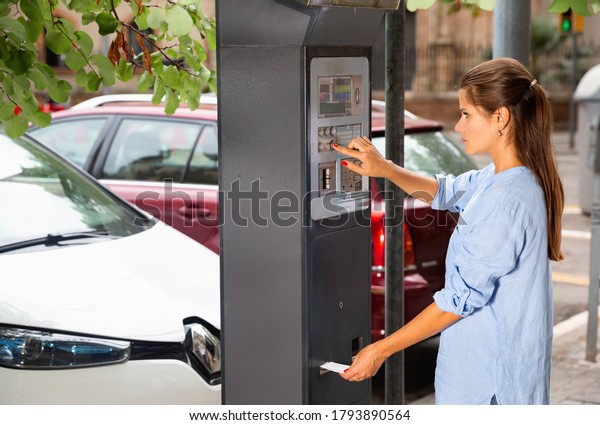 Young positive woman paying for parking in modern\
parking meter on city\
street