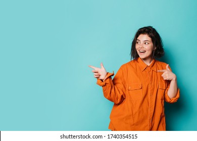 Young Positive Woman Stand On Left Side And Point To Right With Index Fingers. Isolated Over Blue Background. Cheerful Happy Teenager Girl In Orange Shirt Posing On Camera