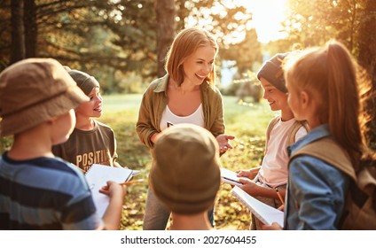 Young positive woman teacher interacting with group of school kids boys and girls during ecology lesson outdoors in forest, kids with notebooks learning about nature and wildlife on sunny autumn day - Powered by Shutterstock