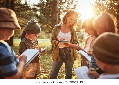 Young Positive Woman Teacher Interacting With Group Of School Kids Boys And Girls During Ecology Lesson Outdoors In Forest, Kids With Notebooks Learning About Nature And Wildlife On Sunny Autumn Day