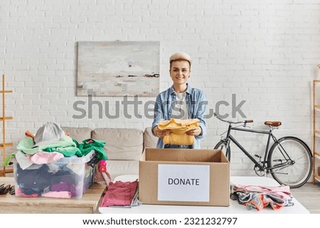 young and positive woman with trendy hairstyle smiling at camera and holding yellow jumper near donation box and plastic container with clothes, sustainable living and social responsibility concept