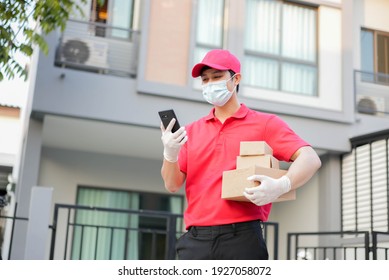 Young Postal Worker Wear Mask And Glove With Red Uniform Carry Box And Stand In Front Of House And Use Smartphone To Call To Customer For Contactless Delivery And E-commerce New Normal In Coronavirus