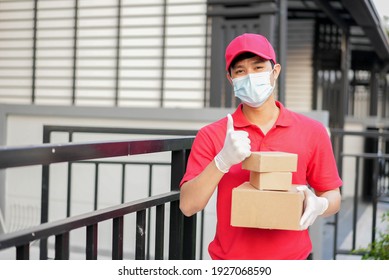 Young Postal Worker Wear Mask And Thumb Up On Glove With Red Uniform Carry Box And Stand In Front Of House To Give Order To Customer For Contactless Delivery And E-commerce New Normal In Coronavirus