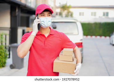 Young Postal Worker Wear Mask And Glove With Red Uniform Carry Box And Stand In Front Of House And Use Smartphone To Call To Customer For Contactless Delivery And E-commerce New Normal In Coronavirus