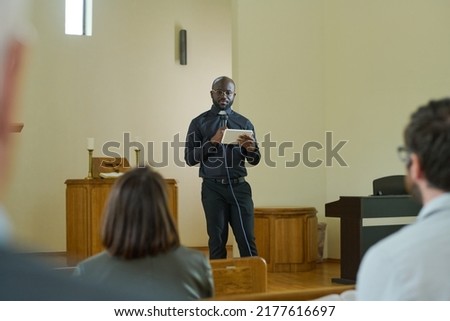 Young preacher in smart casualwear quotating verses from Bible during sermon while standing in front of parishioners and using tablet