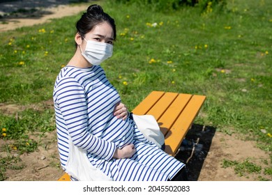 A Young Pregant Asian Woman With A Mask Sits On The Outside Bench And Worries About Her Baby In Her Stomach