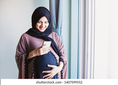 Junge schwangere Arab-Frau in Hijab, die ihr Mobiltelefon benutzt, um Nachrichten zu versenden