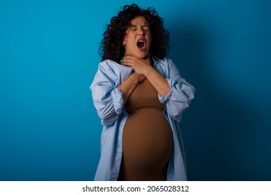 Young pregnant arab woman standing on blue studio background shouting suffocate because painful strangle. Health problem. Asphyxiate and suicide concept.
