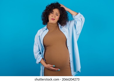 Young pregnant arab woman standing on blue studio background  being confused and wonders about something. Holding hand on head, uncertain with doubt. Pensive concept.