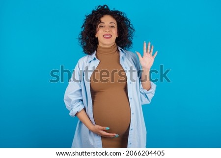 Young pregnant arab woman standing on blue studio background  showing and pointing up with fingers number five while smiling confident and happy.