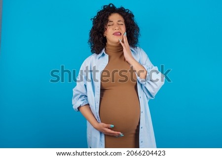 Young pregnant arab woman standing on blue studio background  touching mouth with hand with painful expression because of toothache or dental illness on teeth.