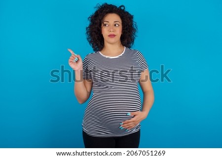 Young pregnant arab woman wearing striped T-shirt on blue studio background points at copy space and advertises something, advices best price.
